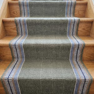 May include: A green stair runner with a patterned border in blue, tan, and white. The runner is installed on wooden stairs. The runner's design features a textured appearance, adding visual interest to the staircase.