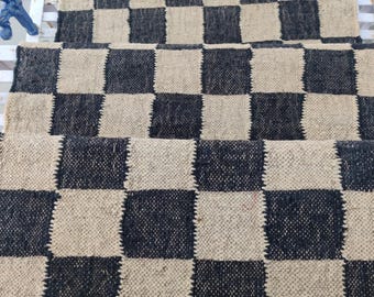 May include: Black and white checkered rug with a woven texture. The rug is made of natural fibers and has a rustic look. It is perfect for adding a touch of style to any room.