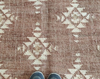 May include: A woven rug with a repeating geometric pattern in shades of brown and cream. The rug's texture is visible, and a pair of blue and gray athletic shoes are at the bottom of the image.