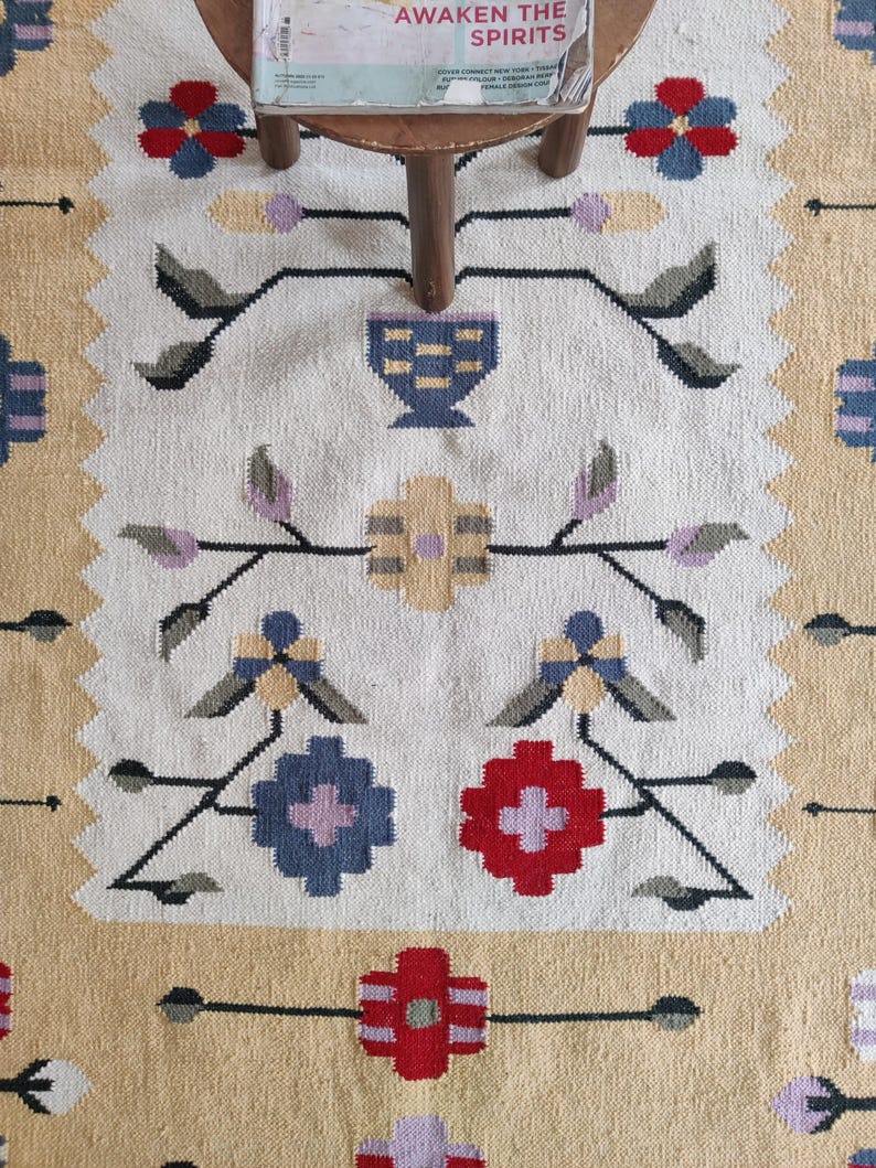 Puede incluir: Una alfombra tejida con un centro de color crema y un patr&oacute;n repetitivo de flores estilizadas y formas geom&eacute;tricas en rojo, azul, amarillo y gris. La alfombra tiene un borde ancho de color tostado y un dise&ntilde;o dentado en la secci&oacute;n interior.