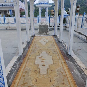 May include: A beige and cream patterned rug with a white fringe. The rug is laid out in a white building with a white archway and blue trim.