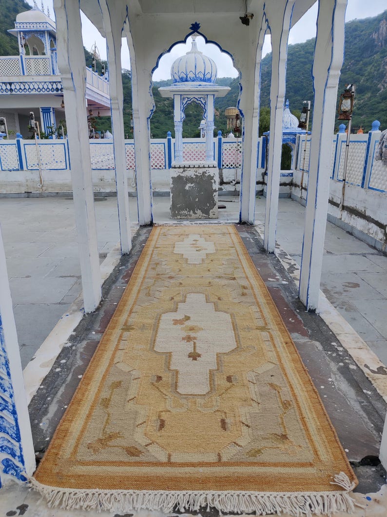 May include: A beige and cream patterned rug with a white fringe. The rug is laid out in a white building with a white archway and blue trim.