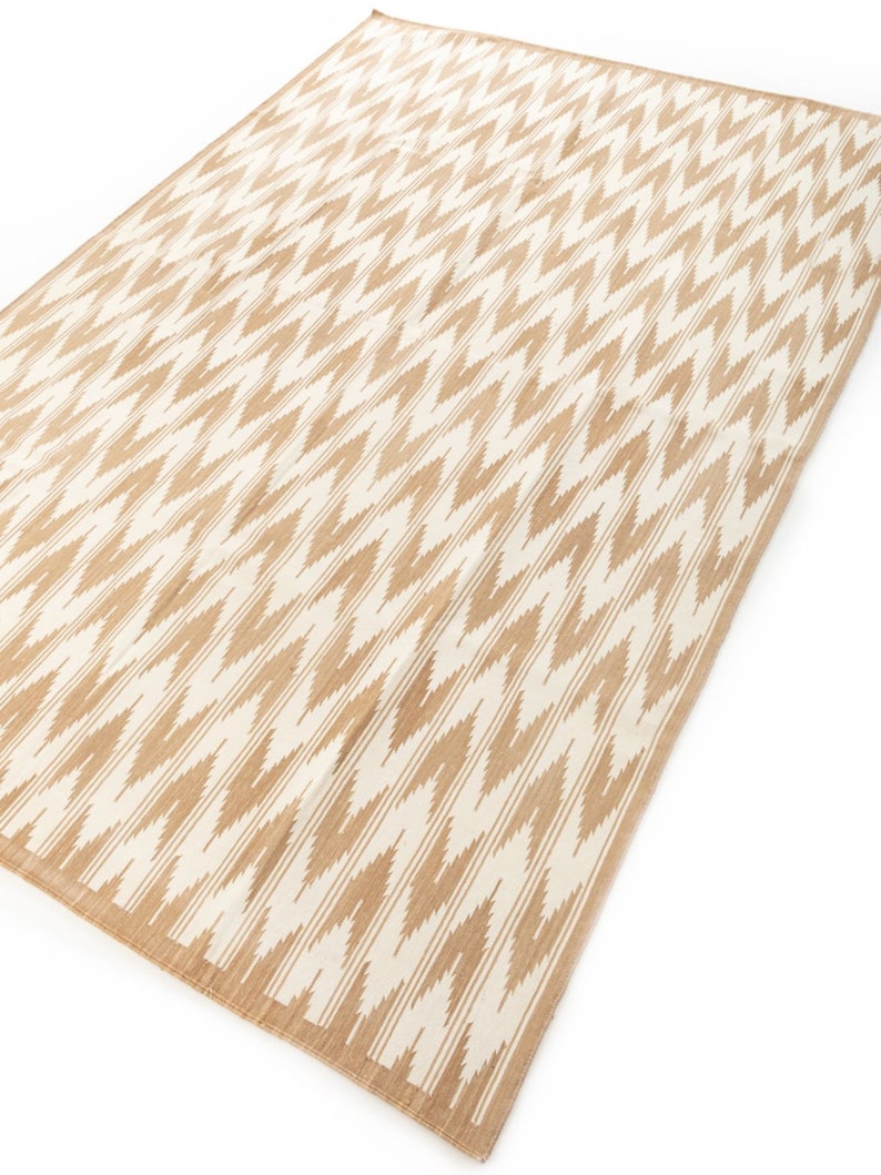 Puede incluir: Una alfombra beige y blanca con un dise&ntilde;o de chevron. La alfombra est&aacute; hecha de un material tejido y tiene un brillo sutil.
