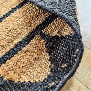 May include: Close-up of a woven jute rug with black stripes. The rug has a natural, earthy texture and is made of natural fibers.
