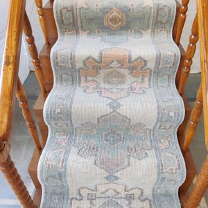 May include: A light blue and beige patterned rug runner with a geometric design. The rug is on a wooden staircase with a wooden handrail.