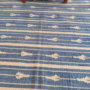 Op de afbeelding: Blauw-wit gestreept tapijt met een subtiel patroon van witte bloemen. Het tapijt is gemaakt van geweven materiaal en heeft een zachte, textuur.