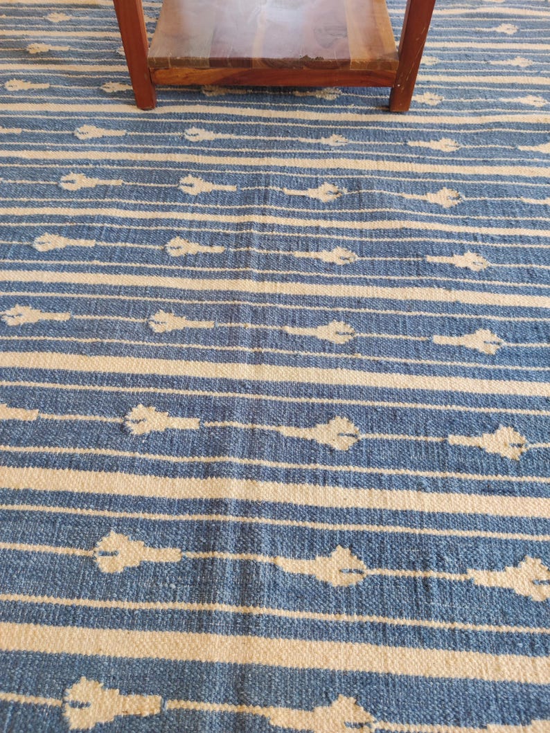 Op de afbeelding: Blauw-wit gestreept tapijt met een subtiel patroon van witte bloemen. Het tapijt is gemaakt van geweven materiaal en heeft een zachte, textuur.