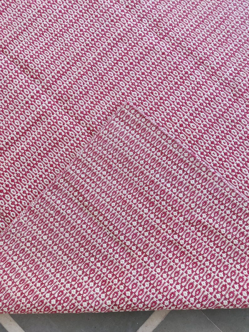 May include: A close-up of a woven textile with a repeating diamond pattern. The fabric is primarily a deep red color with a contrasting off-white pattern. The texture appears soft and the weave is tight.