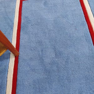 May include: A light blue rug with a rectangular design outlined in red and white. The rug has a soft, textured surface and a simple, modern aesthetic. The corner of a wooden table leg is visible.