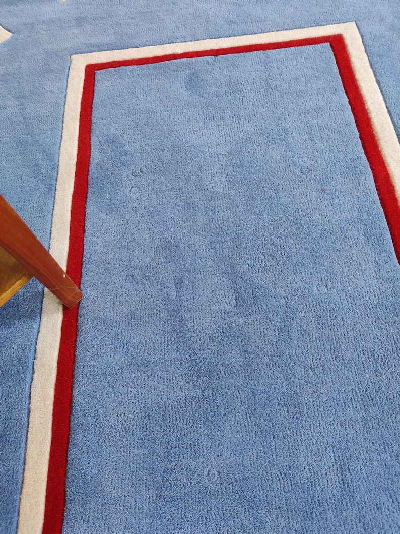 May include: A light blue rug with a rectangular design outlined in red and white. The rug has a soft, textured surface and a simple, modern aesthetic. The corner of a wooden table leg is visible.
