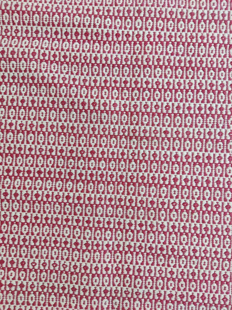 May include: Close-up of a woven textile with a repeating pattern. The design features a series of small, white, diamond-shaped motifs set against a red background. The fabric has a textured appearance, suggesting a natural fiber.
