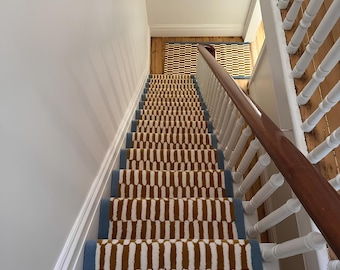 Puede incluir: Escalera con una alfombra con estampado. La alfombra presenta un diseño repetitivo de formas verticales marrones y blancas, bordeado por un borde azul. La escalera tiene una barandilla blanca y un pasamanos de madera.