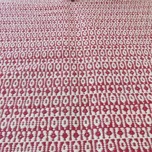 May include: A close-up of a red and white patterned rug. The rug has a geometric design with repeating diamond and oval shapes. The rug is made of woven material and has a textured surface. The legs of a wooden chair are visible in the top of the image.