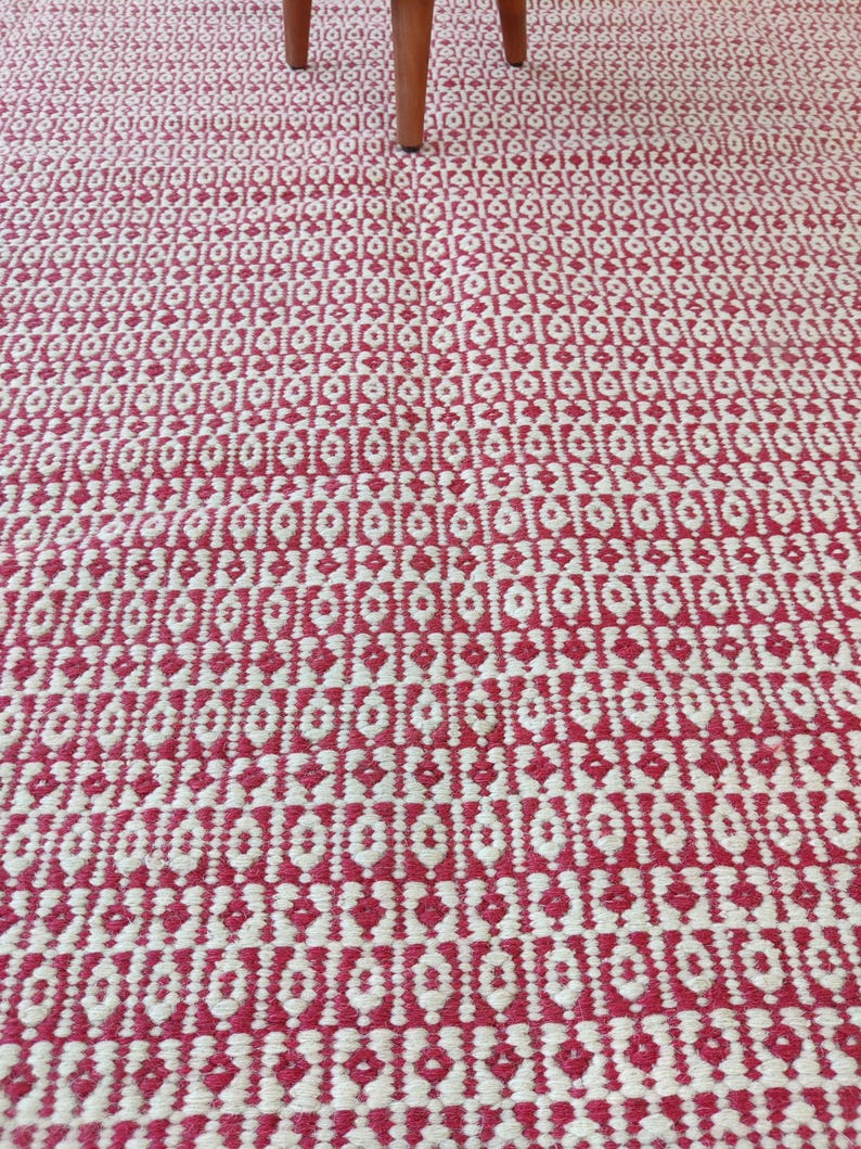May include: A close-up of a red and white patterned rug. The rug has a geometric design with repeating diamond and oval shapes. The rug is made of woven material and has a textured surface. The legs of a wooden chair are visible in the top of the image.