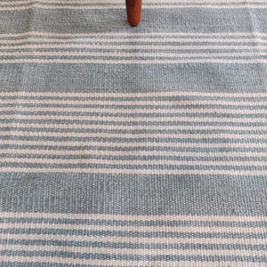 Pu&ograve; includere: Primo piano di un tappeto a righe blu e bianche con una gamba di un mobile in legno. Il tappeto presenta strisce orizzontali azzurre e bianche. La gamba in legno &egrave; di colore marrone scuro.