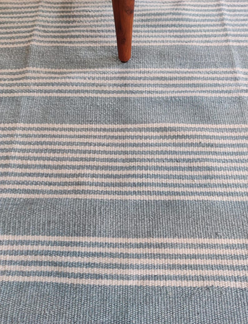 Pu&ograve; includere: Primo piano di un tappeto a righe blu e bianche con una gamba di un mobile in legno. Il tappeto presenta strisce orizzontali azzurre e bianche. La gamba in legno &egrave; di colore marrone scuro.