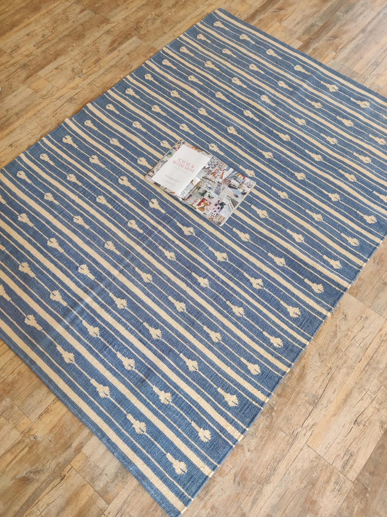 Op de afbeelding: Een blauw en wit gestreept tapijt met een subtiel bloemenpatroon. Het tapijt is gemaakt van katoen en is perfect om elke kamer een vleugje stijl te geven.