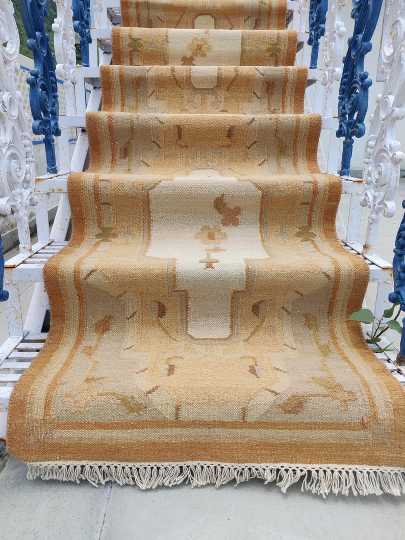 May include: A beige and brown patterned rug with fringe on the bottom edge. The rug is laid out on a set of white stairs with a blue metal railing.