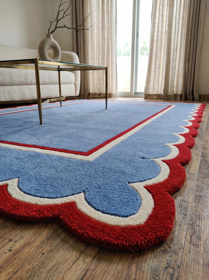 May include: A blue area rug with a scalloped red border and a white and red inner border. The rug is placed on a wooden floor, with a glass-topped coffee table and a white sofa in the background. The rug's design adds a decorative element to the room.