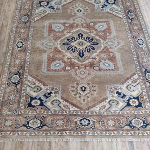 May include: A brown rug with a beige and blue patterned design. The rug has a fringe border.