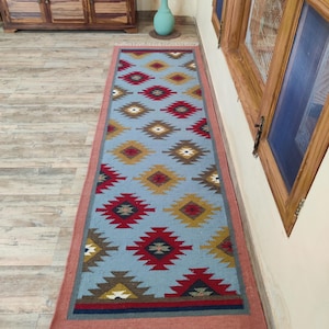 Può includere: Un tappeto rettangolare azzurro con un motivo geometrico rosso, giallo, marrone e bianco. Il tappeto ha un bordo rosa e frange bianche alle estremità corte. Il tappeto è posato su un pavimento in legno.