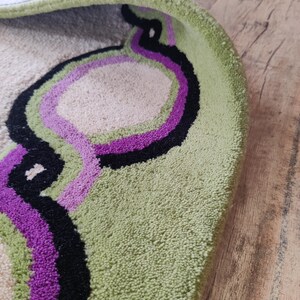 May include: A round rug with a light green border and a beige center. The rug features a design of overlapping, curved lines in black and purple. The rug is on a wooden floor.
