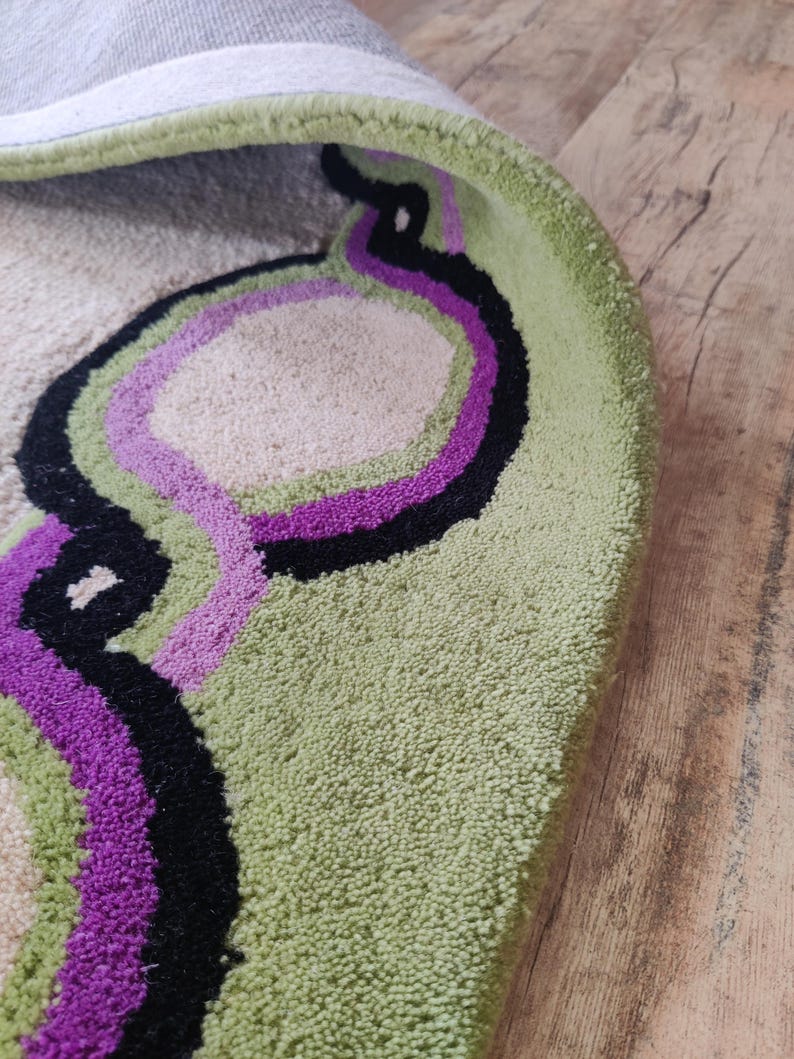 May include: A round rug with a light green border and a beige center. The rug features a design of overlapping, curved lines in black and purple. The rug is on a wooden floor.