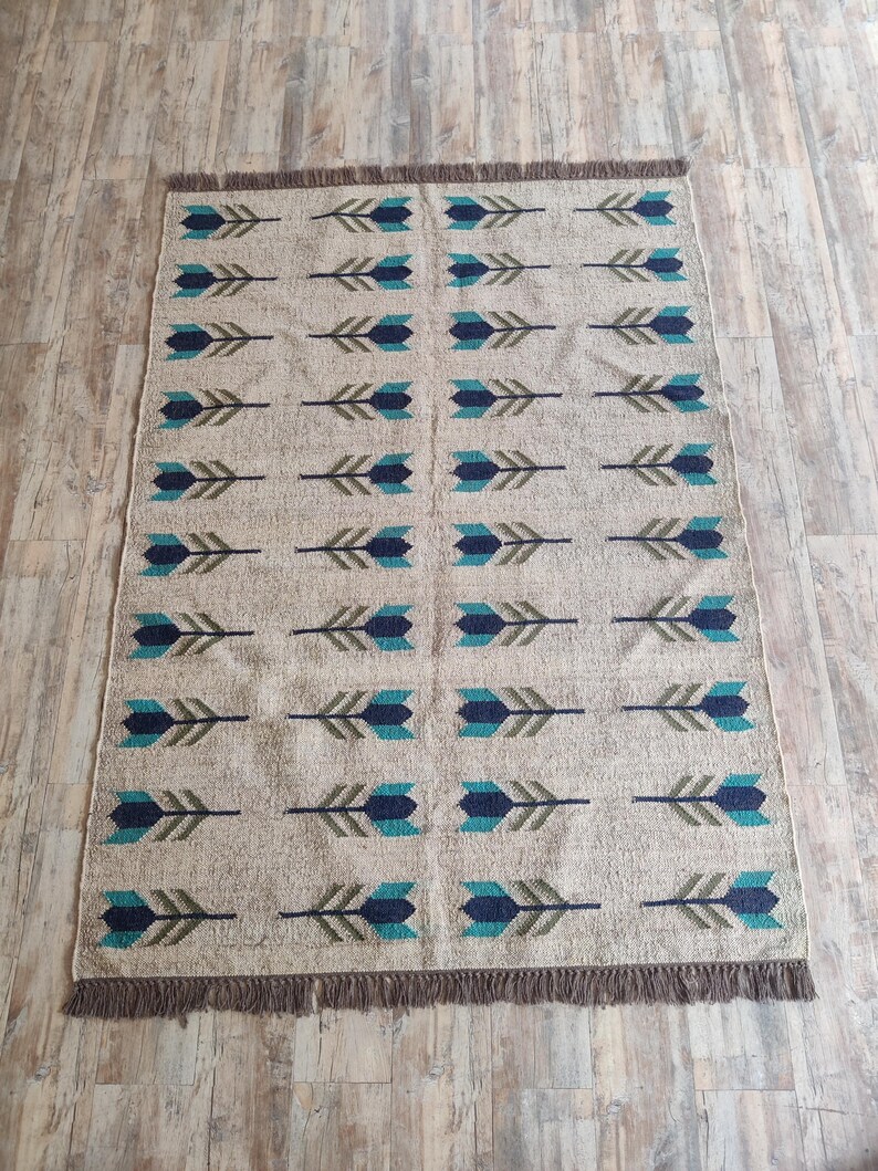 May include: A beige rug with a repeating pattern of blue and green arrows. The rug has a fringe border.