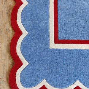 May include: A blue rug with a scalloped edge in red and white. The rug has a rectangular design with a red border and a white inner border. The rug is made of a soft, plush material.