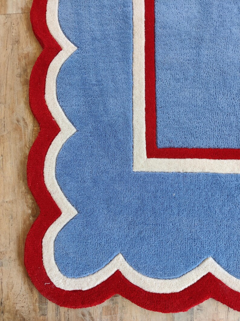 May include: A blue rug with a scalloped edge in red and white. The rug has a rectangular design with a red border and a white inner border. The rug is made of a soft, plush material.