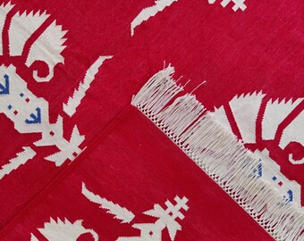 May include: A red rug with a white floral pattern and blue accents. The rug has a fringed edge and a folded corner, revealing the underside. The design is repeated across the surface, creating a bold and eye-catching textile.