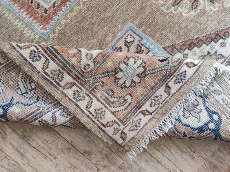 May include: A close-up of a beige and brown patterned rug with a fringe border. The rug is folded in half, showing the intricate details of the design.