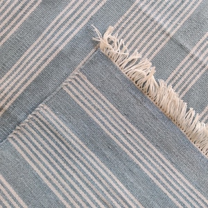 Pu&ograve; includere: Un tappeto a righe blu e bianche con un bordo a frange. Il tappeto presenta strisce alternate blu chiaro e bianche, creando un motivo strutturato classico. L'angolo del tappeto &egrave; ripiegato, rivelando il lato inferiore.