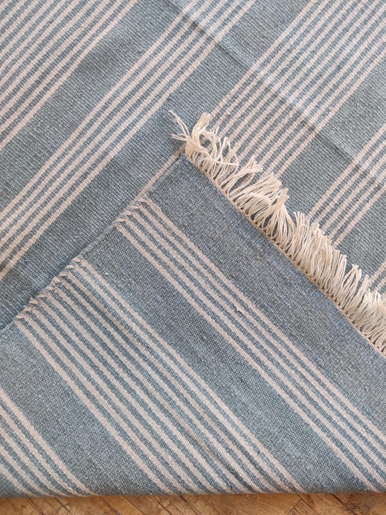Pu&ograve; includere: Un tappeto a righe blu e bianche con un bordo a frange. Il tappeto presenta strisce alternate blu chiaro e bianche, creando un motivo strutturato classico. L'angolo del tappeto &egrave; ripiegato, rivelando il lato inferiore.