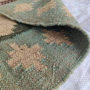 May include: A close-up of a green and beige woven rug with a geometric pattern. The rug is made of natural fibers and has a fringed edge.