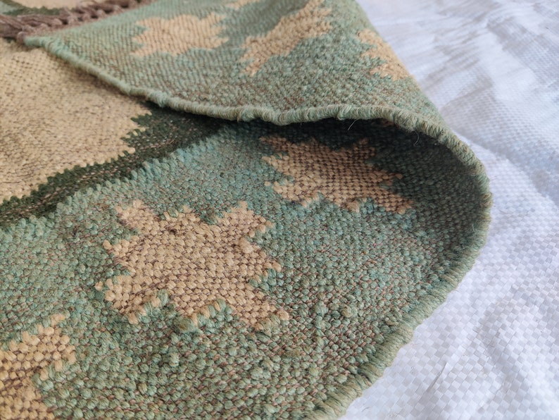 May include: A close-up of a green and beige woven rug with a geometric pattern. The rug is made of natural fibers and has a fringed edge.