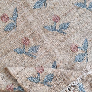 Può includere: Tappeto beige tessuto con motivo floreale. Il tappeto presenta disegni ripetuti di fiori rosa e foglie blu. Il tappeto è ripiegato, rivelando un bordo sfrangiato. Il tappeto è realizzato in fibra naturale.