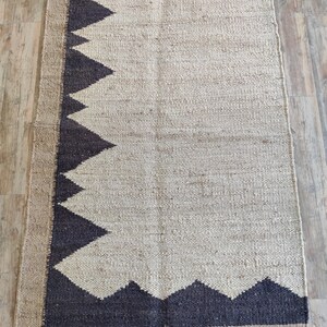 K&ouml;nnte beinhalten: Ein rechteckiger Jute-Teppich mit einem geometrischen Muster in Schwarz und Wei&szlig;. Der Teppich hat einen Fransenrand.