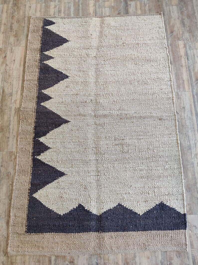 K&ouml;nnte beinhalten: Ein rechteckiger Jute-Teppich mit einem geometrischen Muster in Schwarz und Wei&szlig;. Der Teppich hat einen Fransenrand.