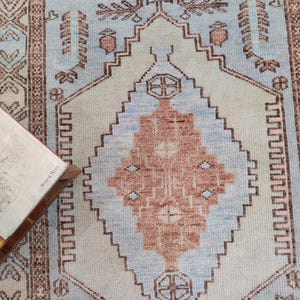 Peut inclure: Un tapis vintage avec un motif géométrique bleu clair et brun. Le tapis a un aspect délavé et vieilli et présente une forme de diamant centrale avec un cercle plus petit à l'intérieur.