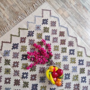 May include: A white rug with a geometric pattern in shades of green, blue, and brown. The rug has a fringe border and features a bowl of fruit in the center.