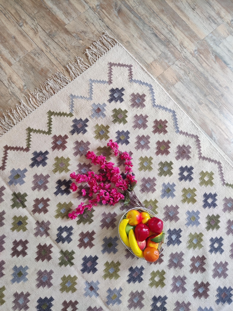 May include: A white rug with a geometric pattern in shades of green, blue, and brown. The rug has a fringe border and features a bowl of fruit in the center.