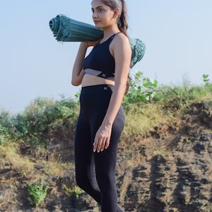 May include: A woman in black athletic wear carries a rolled-up green yoga mat over her shoulder. She is walking barefoot on a dirt path.