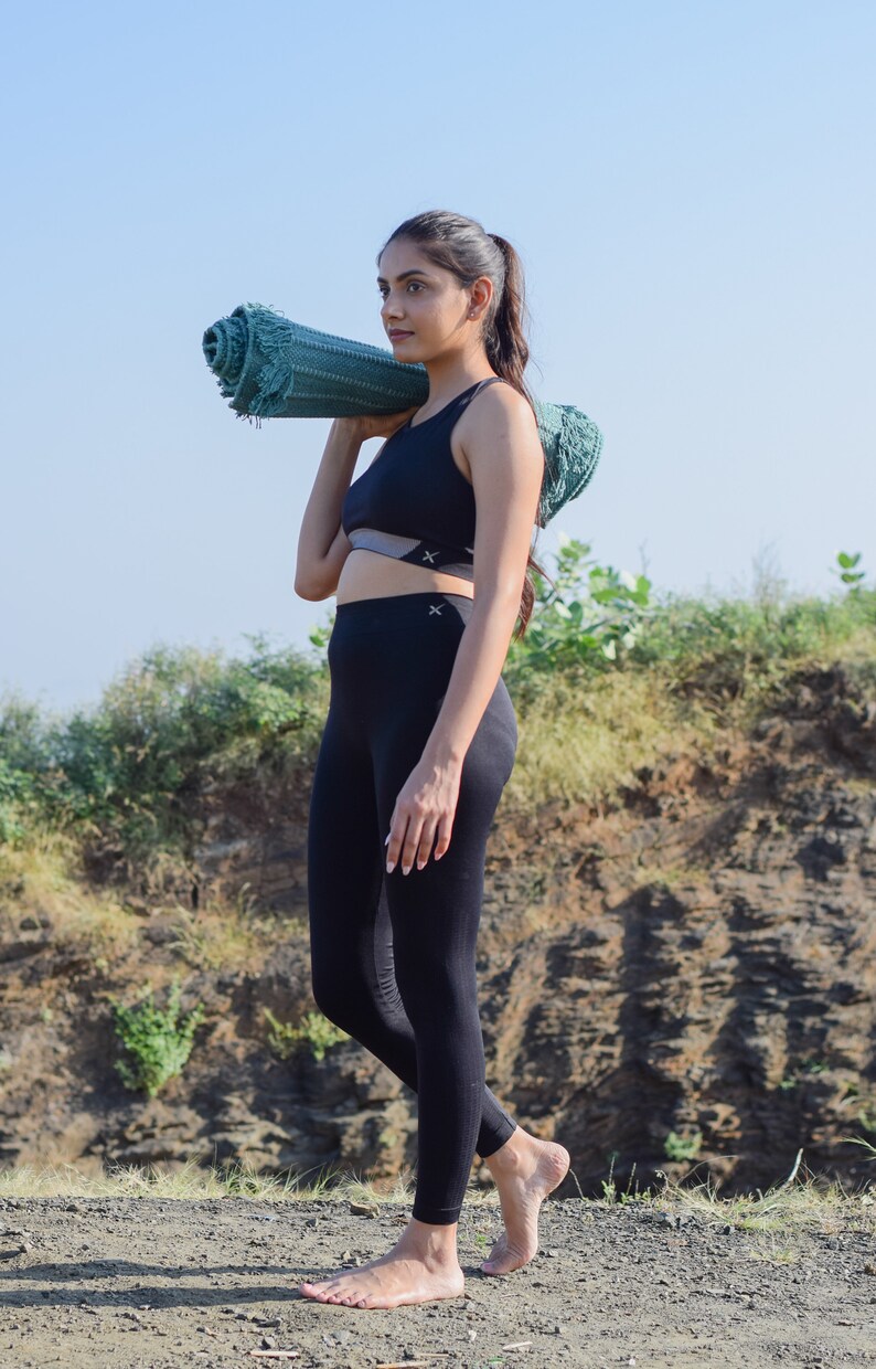 May include: A woman in black athletic wear carries a rolled-up green yoga mat over her shoulder. She is walking barefoot on a dirt path.