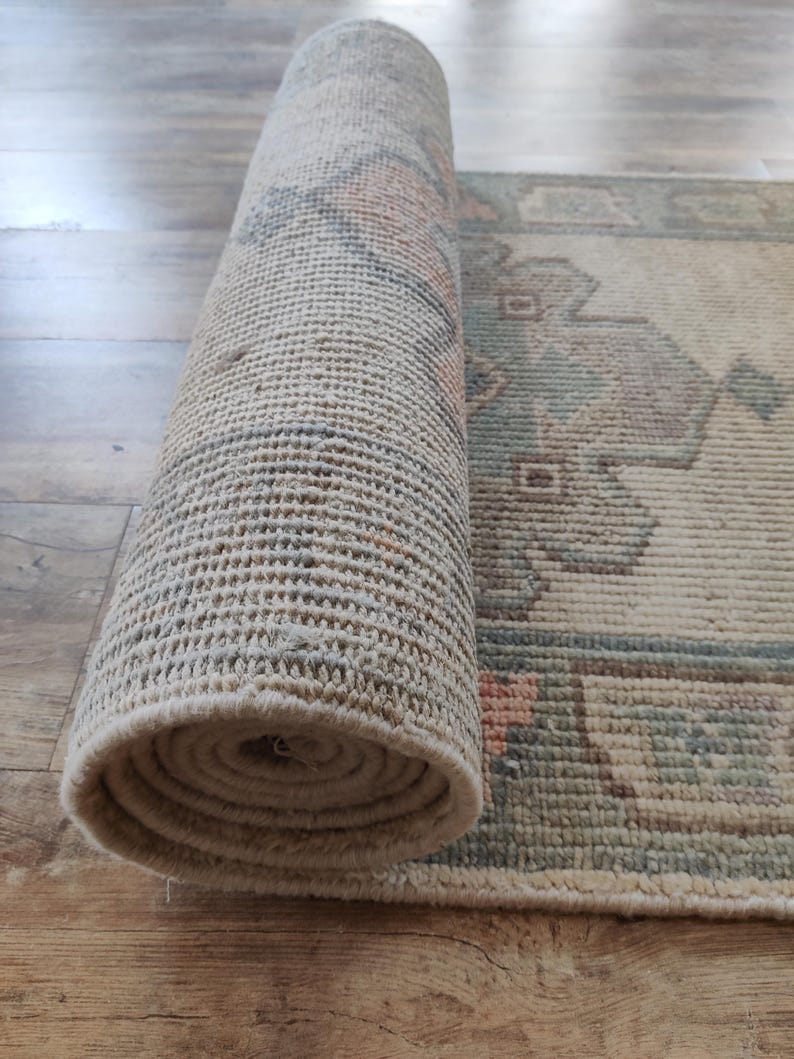 May include: A rolled up beige rug with a geometric pattern in shades of blue, green, and pink. The rug is woven with a textured surface.