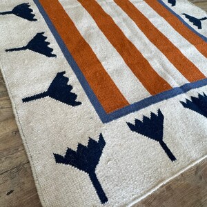 May include: A white rug with orange and blue stripes and a border of blue stylized flowers.