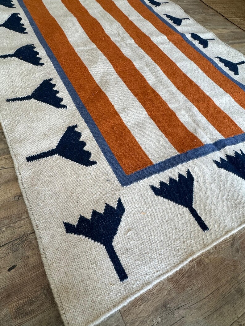 May include: A white rug with orange and blue stripes and a border of blue stylized flowers.