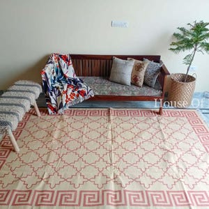 Pode incluir: Um tapete de cor creme com um padrão geométrico vermelho. O tapete está colocado sobre um piso de madeira em uma sala de estar. Um banco de madeira com um almofada listrada branca e cinza está em primeiro plano. Um sofá de madeira com almofadas e uma manta de arremesso floral está no fundo. O texto "House of Kilims" é visível no canto inferior direito da imagem.