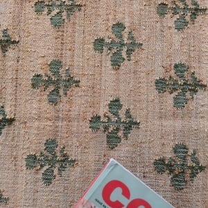 Puede incluir: Primer plano de una tela beige con un patrón repetido de diseños de trébol de cuatro hojas verdes. La tela tiene una apariencia texturizada y tejida.