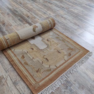 May include: A rolled up beige and brown rug with a geometric pattern and white fringe. The rug is lying on a light wood floor.